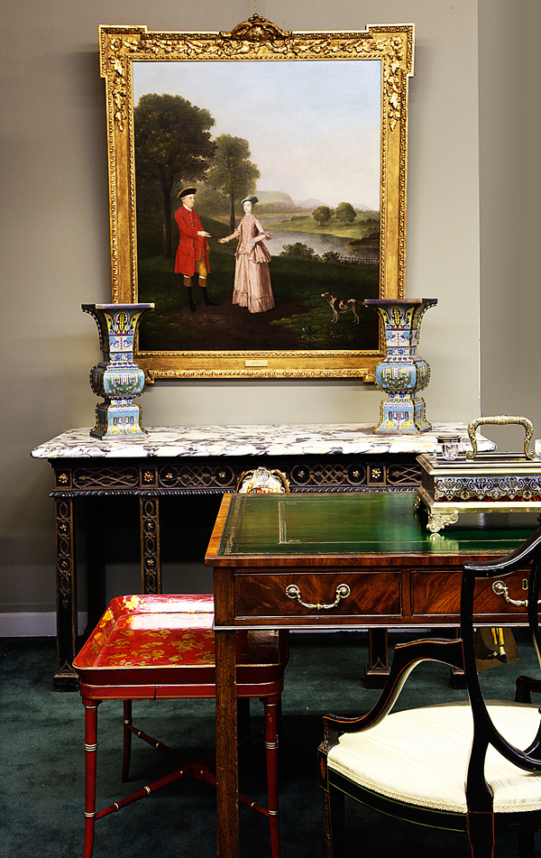 A portrait by 18th-century English painter Arthur Devis hangs over a George II ebonized-and-gilt-marble-top console table that commemorates the marriage of Thomas Pelham to Anne Frankland in 1754 and bears the arms of both families; in the foreground is a Regency writing table in mahogany, ca. 1785, and a Victorian gilt papier-mâché tray, ca. 1850.