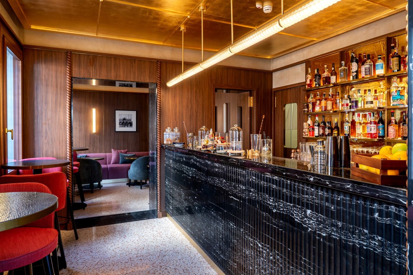 Bar du nouvel hôtel Ca' di Dio, à Venise, avec sol en terrazzo, devanture du comptoir en marbre noir et chaises tapissées de rouge autour des tables rondes. Architecture et décoration intérieure de l'hôtel réalisées par Patricia Urquiola