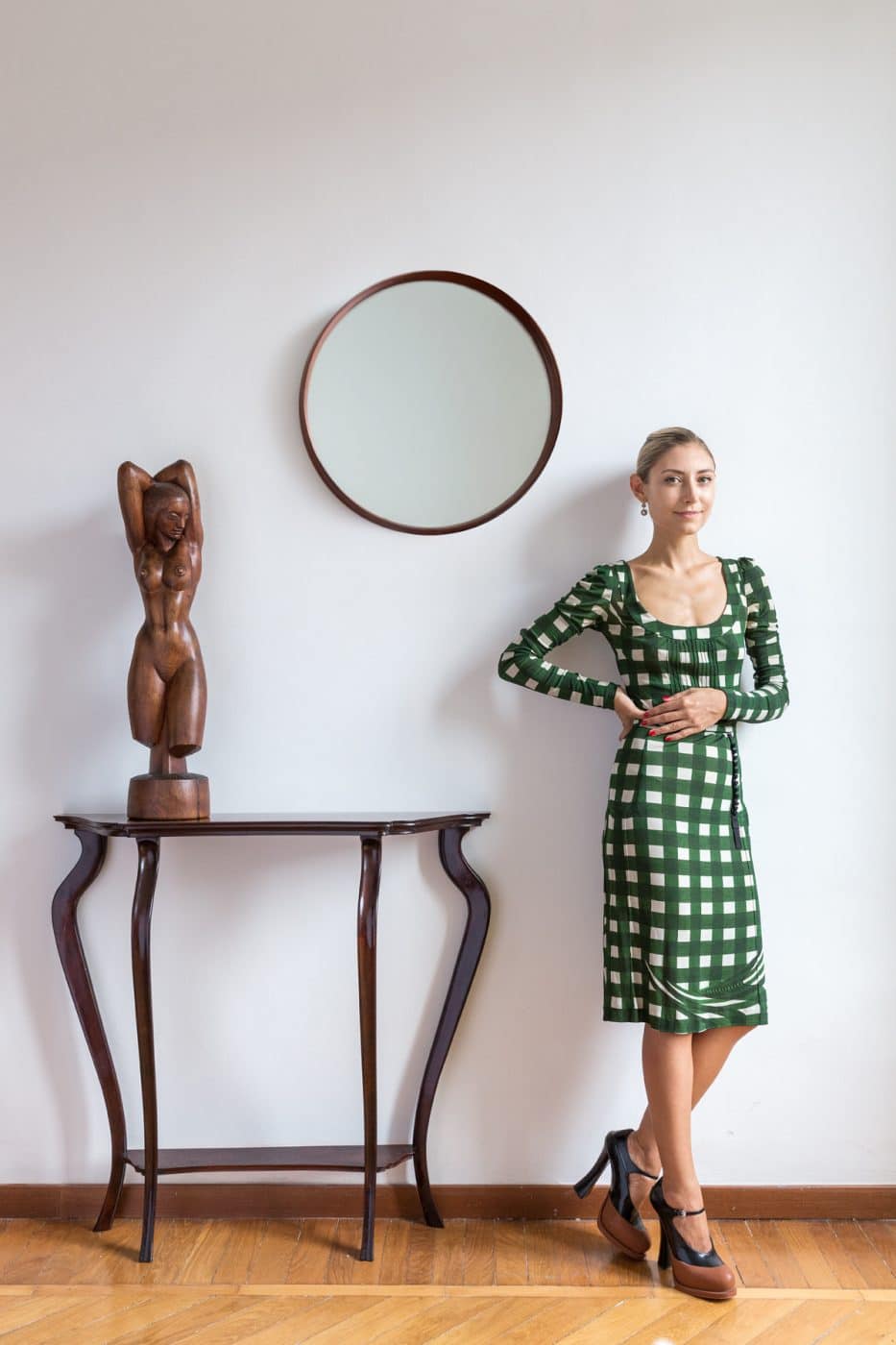 Walton in einem grün karierten Prada-Kleid neben einem runden holzgerahmten Spiegel, der über einem italienischen Konsolentisch aus der Mitte des Jahrhunderts mit der Holzfigur einer nackten Frau hängt