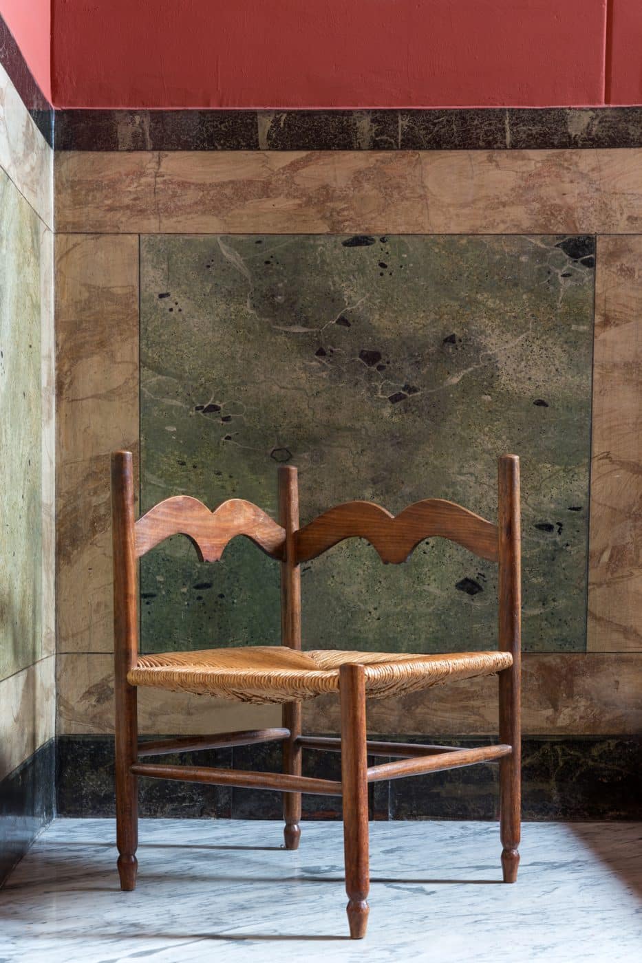 Französischer Eckstuhl aus Eiche mit Sitzfläche aus Binsen, fotografiert vor der farbigen Steinwand im Eingangsbereich des Mailänder Wohnhauses, in dem Jenny Walton lebt
