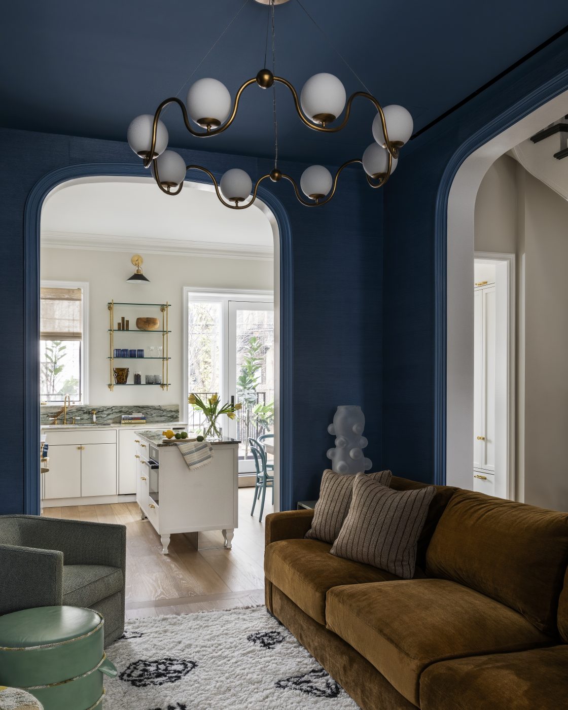 Le salon confortable a des murs bleu foncé et des étagères encastrées, une paire de chaises A. Rudin à dossier en tonneau, deux poufs Nickey Kehoe, un tapis Nordic Knots à motifs de tatouages, un lustre Currey and Company aux verres ondulés et un canapé Kravet.